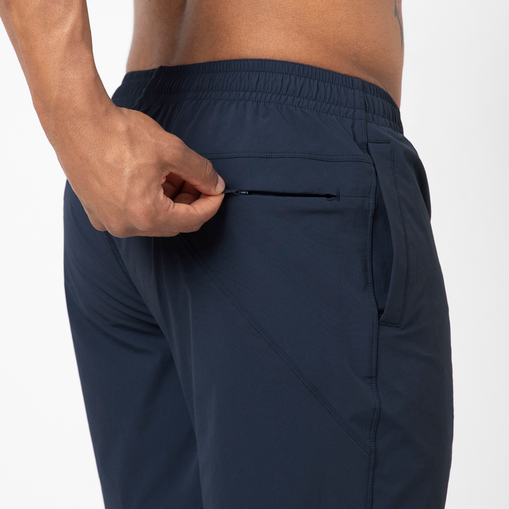 A person wearing Atlas Short athletic pants in navy blue with moisture-wicking fabric is unzipping a small side pocket. Only their lower torso and hand are shown against a white background.